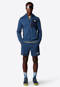 Heren sportoutfit bestaande uit een marineblauwe hoodie met rits en een zak, bijpassende shorts, gradiënt blauwe sokken en grijs-gele sneakers.