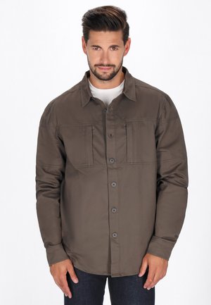 Cămașă maro cu nasturi și guler, cu două buzunare pe piept și mâneci lungi, realizată dintr-un material neted și mat. Pur tată peste un tricou alb.