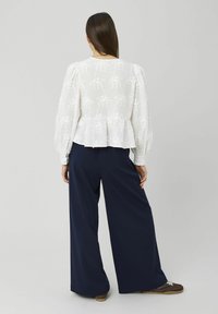 Femme aux longs cheveux bruns portant un chemisier blanc en dentelle florale avec des manches bouffantes et un pantalon large bleu marine, debout de dos.