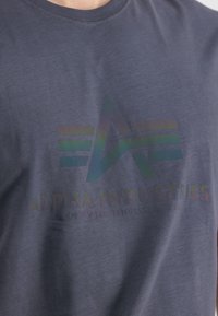 Marineblå bomulds-t-shirt med et blankt iriserende logo og teksten "ALPHA INDUSTRIES, KNOXVILLE, TENNESSEE, USA" på forsiden.