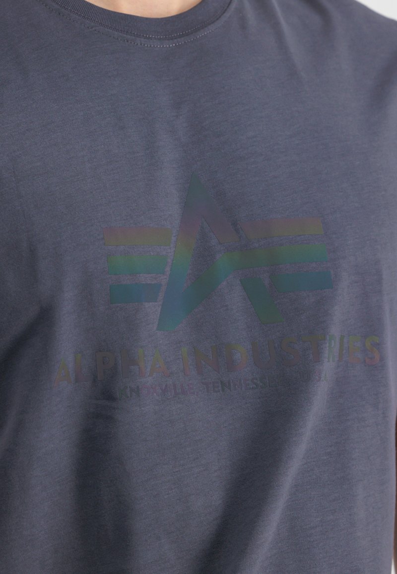 Marineblå bomulds-t-shirt med et blankt iriserende logo og teksten "ALPHA INDUSTRIES, KNOXVILLE, TENNESSEE, USA" på forsiden.