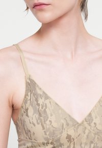 Mulher a usar uma camisola sem mangas com padrão bege e taupe, com finas alças nos ombros, a revelar o decote e a parte superior do peito.