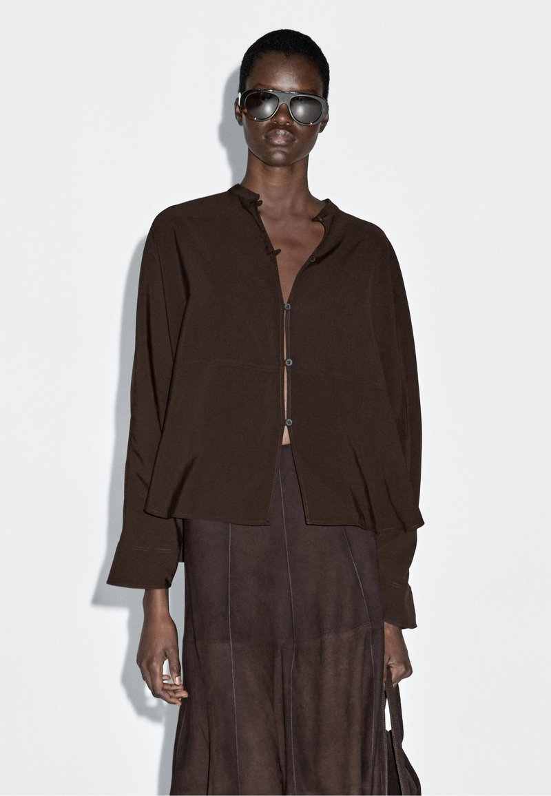 Brązowa koszula na guziki o luźnym kroju, z długimi rękawami i minimalistycznym wzornictwem. Noszona na dopasowaną brązową spódnicę, z delikatnymi przeszyciami.