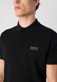 Polo noir en tissu texturé. Présente un col à deux boutons et un petit logo brodé "Barbour International" sur la poitrine.