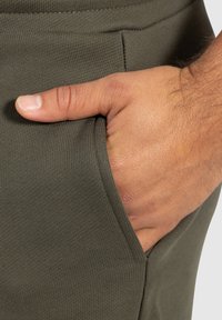 Pantalón chándal verde oliva con textura acanalada, mano descansando dentro del bolsillo. La tela muestra un acabado suave y un detalle fino.