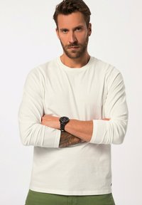 T-shirt bianca a maniche lunghe realizzata in cotone, con scollatura a girocollo e vestibilità rilassata, abbinata a pantaloni verdi. Un orologio nero adorna il polso.