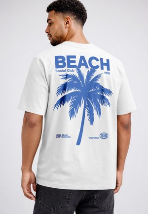 Man facing away wearing white T-shirt with large blue palm tree and text "BEACH Social Club 1976" on back.
