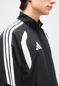 Νεαρό άτομο που φοράει μαύρο αθλητικό μπουφάν με φερμουάρ, με λευκές ρίγες στα μανίκια και λευκό τριγωνικό λογότυπο στο στήθος.