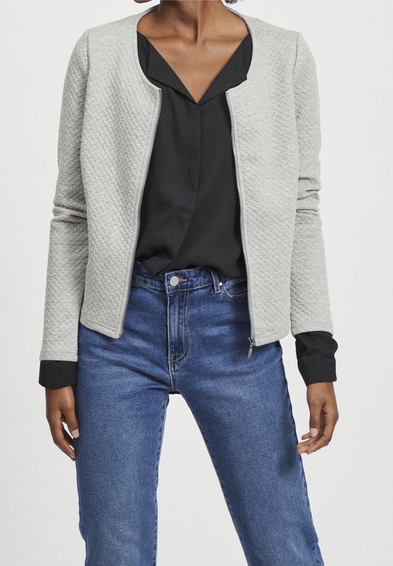 Light gray textured zip-up jacket with a round neckline, worn over a black blouse and paired with high-waisted blue jeans. Dark cuffs visible.