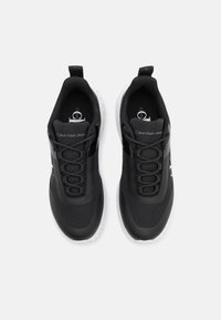 Svarta sportskor med en texturerad ovandel, rund tå och vit sula. Har snörning och logotyp på sidan och tungan.