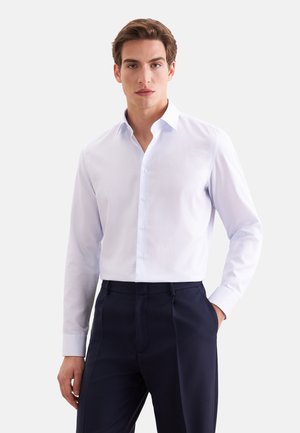 Jeune homme portant une chemise blanche à manches longues boutonnée et un pantalon foncé, debout avec une main dans la poche devant un fond uni.