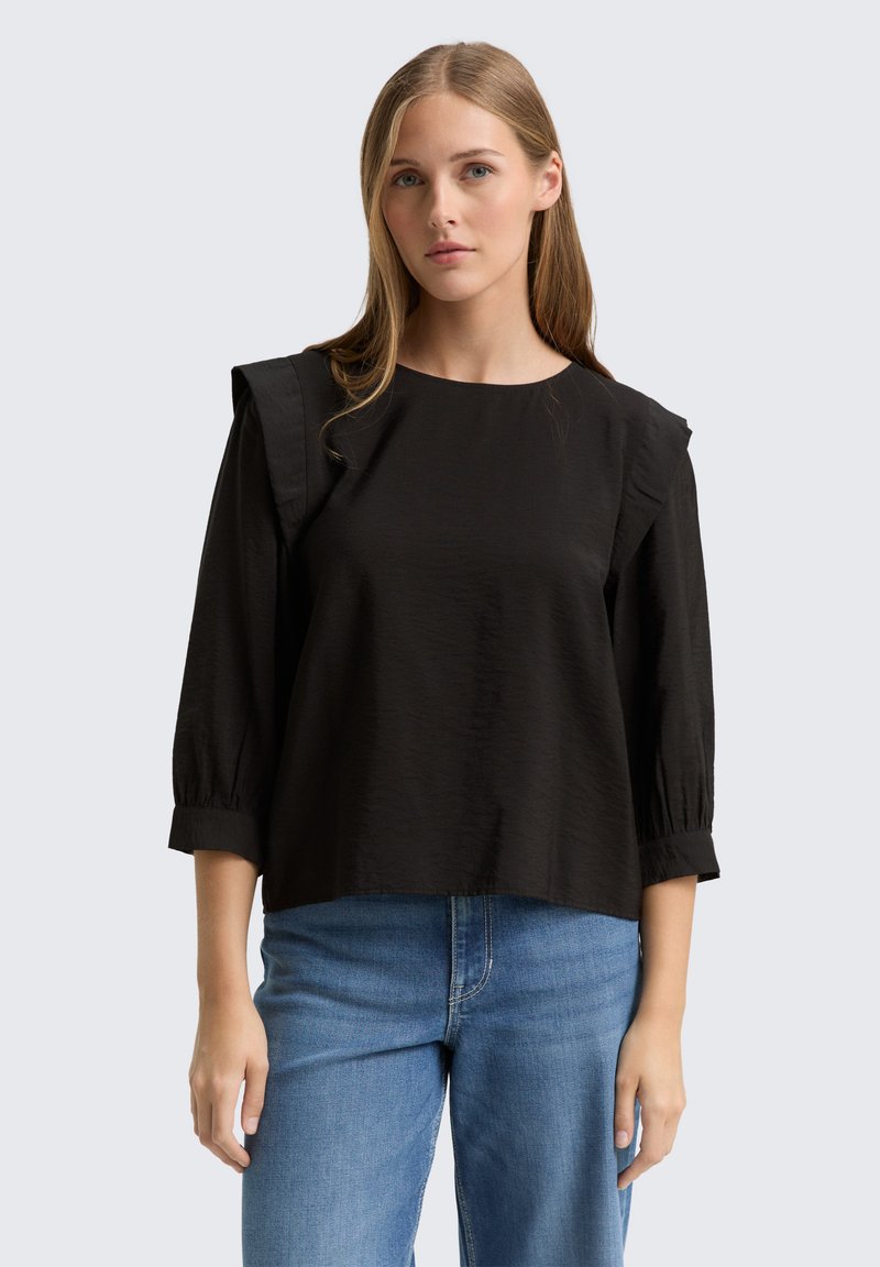 Schwarze Bluse mit kurzen Ärmeln, gefalteten Schulterakzenten und strukturierten Oberflächen, kombiniert mit hellblauen Jeans. Einfaches, lockeres Design.