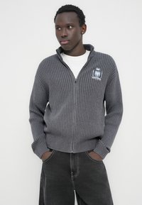 Γκρι ζακάρ πουλόβερ με φερμουάρ και ψηλό γιακά, με κεντημένο λογότυπο στο αριστερό στήθος. Φοριέται πάνω από ένα λευκό T-shirt.