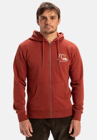 Sweat à capuche rouge avec un zip, poche kangourou, cordon de serrage à la capuche et un logo blanc sur la poitrine gauche. Tissu doux avec une texture lisse.