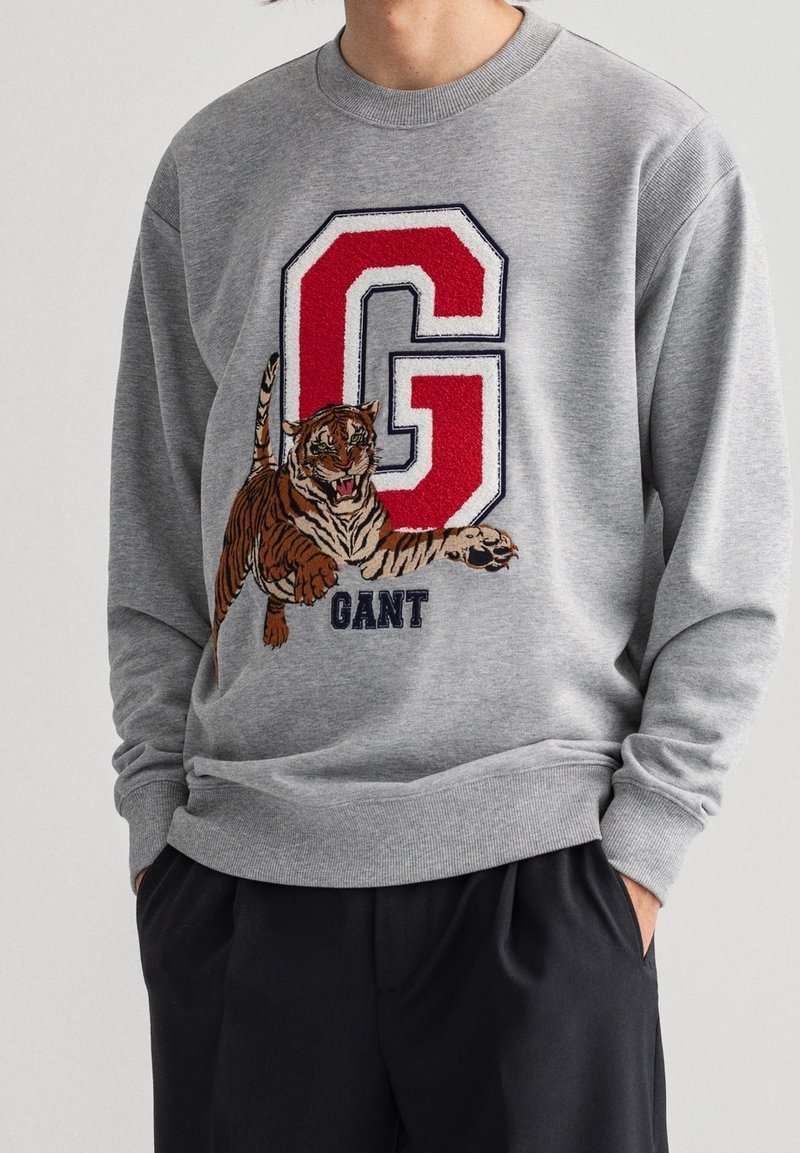 Sudadera gris con una gran letra "G" bordada en rojo y blanco, con un gráfico detallado de un tigre y el texto "GANT" debajo.