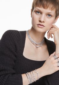 Joyería plateada brillante que incluye collares en capas, pulseras con detalles de corazón y anillos, sobre una blusa negra texturizada.