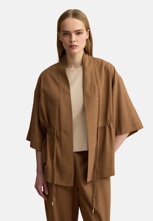 Frau trägt eine lockere braune Jacke mit Kordelzügen über einem beigen Top und passenden braunen Hosen, steht vor einem schlichten Hintergrund.