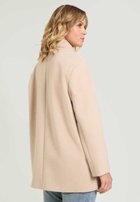 Abrigo de color beige claro con un acabado texturizado. Presenta un corte relajado, mangas largas y un cuello con muescas. La vista trasera muestra líneas limpias y un buen corte.