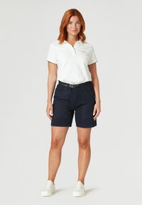 Witte polo met korte mouwen, marineblauwe shorts en witte sneakers. De polo heeft een subtiele logo en een kraag met drie knopen.