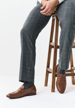 Homme assis sur un tabouret en bois portant un pantalon gris à motifs, des chaussettes foncées et des mocassins en cuir marron avec un détail de bride.