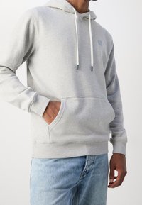 Sudadera gris claro de tela suave, con un bolsillo frontal, cordones con puntas en contraste y un pequeño logo en el pecho.