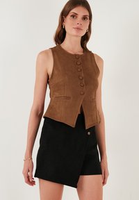 Barna nubuk ujjatlan mellény elöl gombos zárással, két oldalsó zsebbel és testhezálló fazonnal. Fekete nubuk aszimmetrikus szoknya felett viselve.