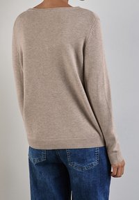 Person som bär en beige stickad tröja och blå denimjeans, sedd bakifrån mot en slät ljusgrå vägg.