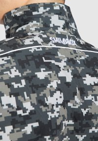 Camouflagejakke i nuancer af grå og sort, med højt krave og "SMILEBOX" broderi i hvidt langs ryggen. Struktureret stofmønster.