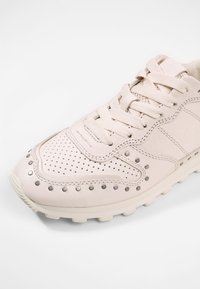 Světle růžové tenisky s perforovaným designem, zaoblenou špičkou, stříbrnými cvočky kolem střední podrážky a plochými tkaničkami pro bezpečné uchycení.