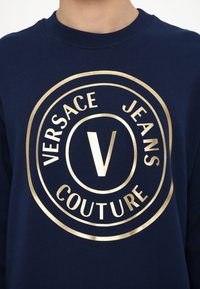 Laivyno spalvos džemperis su apvaliu auksiniu logotipu, kuriame yra "VERSACE JEANS COUTURE" ir išraiškingas "V" centre. Minkšta tekstūra.