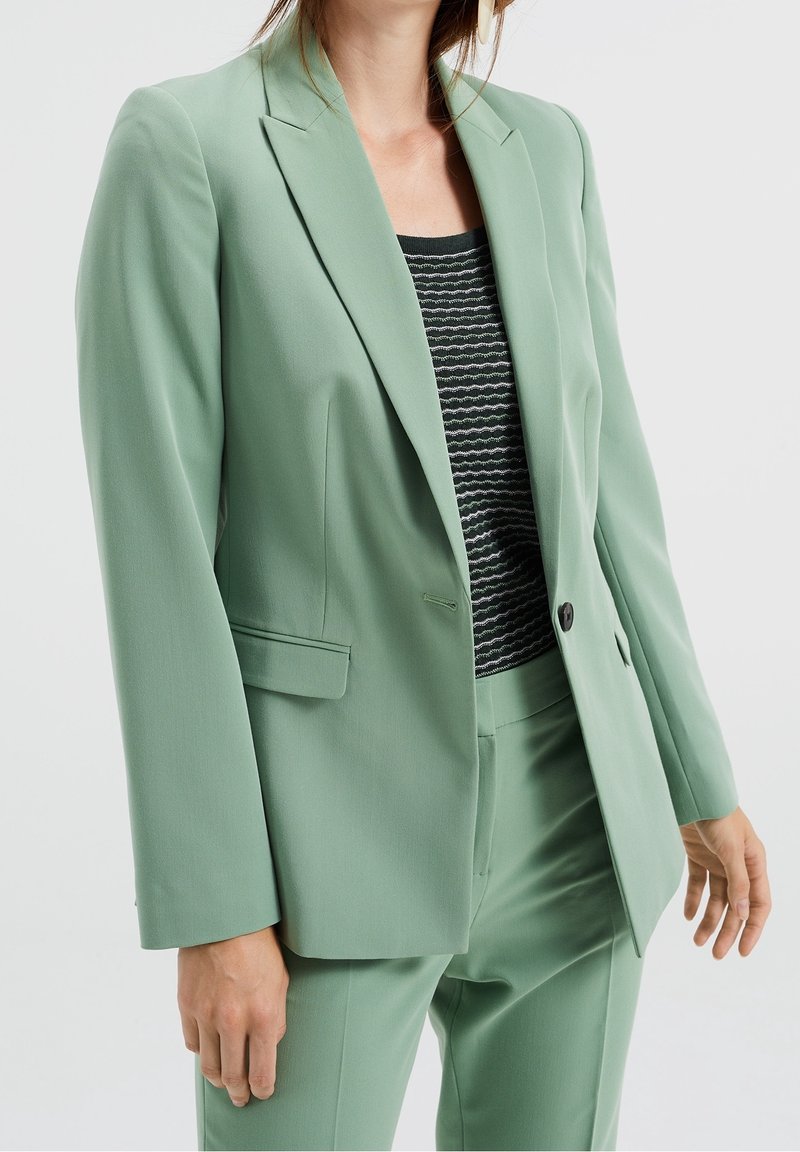 Femme portant un costume vert clair sur mesure avec une veste assortie et un pantalon, par-dessus un haut à motif noir et blanc.