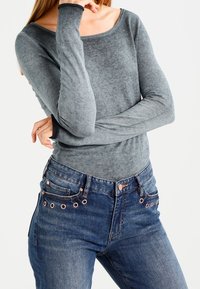 Långärmad, ljusgrå tröja med en vid halsringning, kombinerad med blå denimjeans med roséguldtonade knappar och öglor.