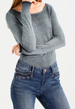 Maglietta a maniche lunghe di un leggero grigio chiaro con un ampio scollo, abbinata a jeans in denim blu con bottoni in acciaio rosa e dettagli a occhiello.
