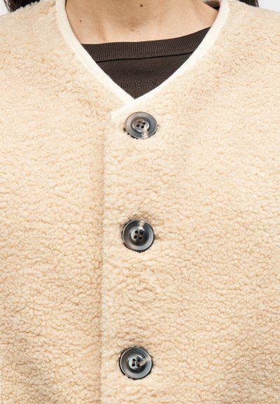 Μπεζ φλις μπουφάν με υφή στην επιφάνεια. Διαθέτει λαιμόκοψη V και πέντε σκοτεινά κουμπιά. Περιμετρικά έχει διακόσμηση από εκρού ύφασμα.