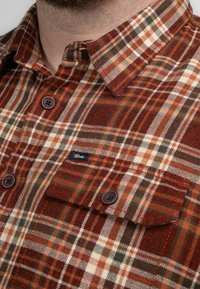 Rood en bruin geruit flanellen overhemd met een button-down kraag, voorzien van bruine knopen en een klein merklabel op de borstzak.