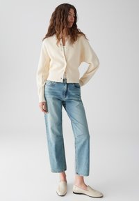 Crème gebreide cardigan met knopen, lichtblauwe hoogwatige jeans met duidelijke stiksels en beige loafers. Gladde texturen overal.