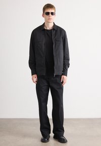 Black zip-up jacket with two chest pockets, paired with a black T-shirt and loose-fitting black pants. Model wears dark sunglasses and shoes.