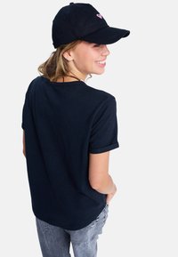 Young woman smiling, wearing a black cap with pink logo, black t-shirt, and light gray jeans, looking over her shoulder against white background.