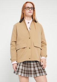 Tαν παλτό με σκούρο γιακά, με κλείσιμο με κουμπιά και δύο μπροστινές τσέπες. Συνδυασμένο με καρό πλισέ φούστα σε αποχρώσεις καφέ και λευκού.