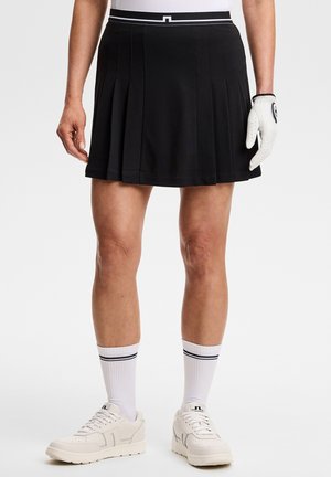 Personne portant une jupe plissée noire, des chaussettes blanches mi-mollet avec des rayures noires, des baskets blanches et un gant de golf blanc sur la main droite.