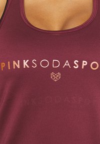 Burgundy linne med rundad halsringning. Har en guld "PINKSODA" logotyp och geometrisk hjärtformad design. Slät textur, sportig passform.
