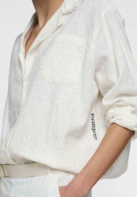 Witte linnen blouse met een knoopsluiting, linkerklepzak, opgerolde mouwen en zwarte bedrukking op de linkermouw.
