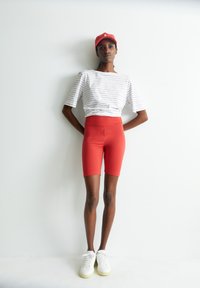 Rode high-waisted fietsshorts gecombineerd met een wit gestreept T-shirt met korte mouwen en een rode pet, afgewerkt met witte sneakers.