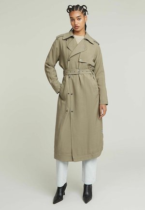 Khaki płaszcz trench z paskiem w talii, dużym kołnierzem i guzikami. Noszony na jasnoniebieskie dżinsy i czarne botki za kostkę.