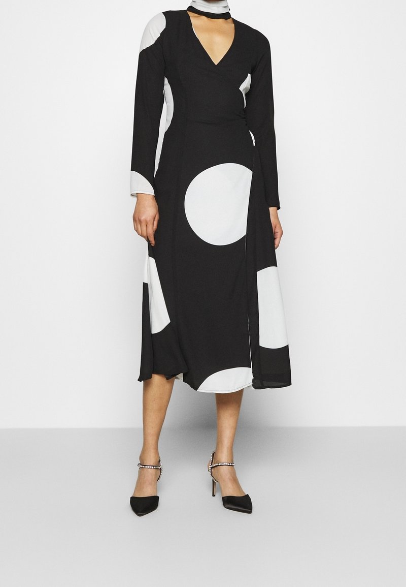 Schwarz-weißes Kleid mit langen Ärmeln, einem V-Ausschnitt und großen kreisförmigen Mustern; kombiniert mit schwarzen High Heels mit Knöchelriemen.