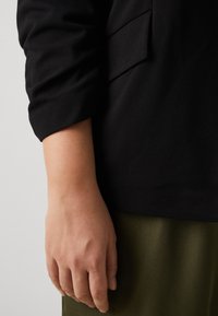 Černý látkový top s přeloženým rukávem a boční kapsou. Ruka spočívá na olivově zelené sukni, která má hladké textury.