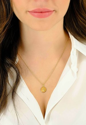 Collier pendentif en or avec un charme circulaire sur une chaîne délicate, porté sur une chemise blanche à col. Texture lisse et finition polie.