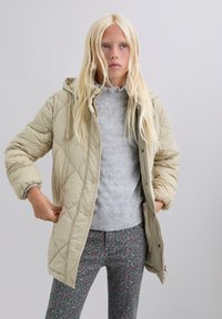Blonde vrouw die een beige gewatteerde capuchonjack over een lichtgrijs gebreid sweater en groene luipaardprint broek aanpast, staande tegen een effen achtergrond.