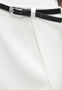 Μαύρη δερμάτινη ζώνη με ασημένια πόρπη δεμένη γύρω από τη μέση λευκών υφασμάτινων παντελονιών με υφή.