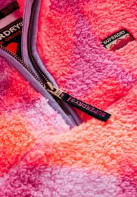 Lähivaade roosale ja oranžile udusetele Superdry jopele, millel on lillakaspunane tõmblukk ja brändinguastmega tõmbluku tõmbaja.